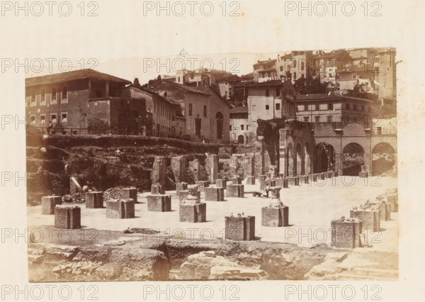 View of the ruins of the Basilica Julia on the Roman Forum in Rome, photograph, page, Roman Forum, c. 1863 - in or before 1873, photographer, Rome, Forum Romanum, paper, albumen print, height 92 mm x width 150 mm, ruin of a building, architecture, Basilica Julia, A sepia-toned photograph showing a scene of ruins and old buildings with an open foreground filled with rectangular stone blocks arranged in rows, some with debris on top, and a few shallow excavated areas. Behind the blocks a raised embankment leads to a row of multi-story buildings with tiled roofs, shuttered windows, and a central structure with a tall arched doorway. To the right a long arcade of arches and columns connects to more masonry and a ladder leaning against one arch. Scattered stones and rough terrain occupy the lower part of the image, and the sky above is pale and untextured.
