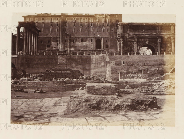 Archaeological remains at the Roman Forum in Rome, photograph, page, Roman Forum, c. 1863 - in or before 1873, photographer, Rome, paper, albumen print, height 99 mm x width 150 mm, landscape with ruins, Forum Romanum, A sepia-toned photograph showing a wide archaeological site with uneven stone pavement in the foreground and rough stone ruins rising toward a higher terrace, scattered blocks and rubble across the site, a low stone wall topped with short cylindrical posts, tall classical columns and column fragments standing on the terrace, a partially preserved archway with ornate stonework on the right, a multi-story building with many rectangular window openings and a plain facade behind the ruins, and a pale border surrounding the image.