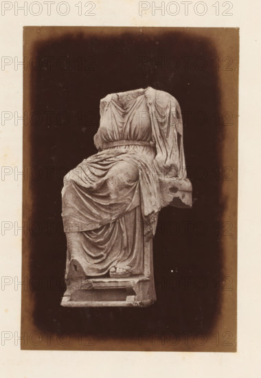 Sculpture on the Palatine Hill, depicting an enthroned Magna Mater, photograph, page, Rome, c. 1863 - in or before 1873, photographer, paper, albumen print, height 150 mm x width 100 mm, sculpture, sitting figure - AA - female human figure, A worn marble statue of a seated draped figure is centered against a dark rectangular background with a lighter border; the figure lacks a head and shows detailed folds of cloth gathered at the waist and cascading over the legs and seat, with one visible foot wearing a sandal resting on a block and parts of the statue showing chips and weathering.