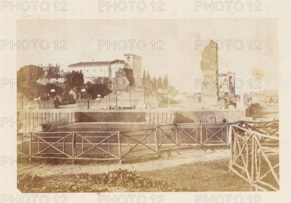 View of ruins (presumably) at Rome, photograph, page, Rome, c. 1863 - in or before 1873, photographer, paper, albumen print, height 100 mm x width 140 mm, landscape with ruins, A sepia-toned view of an open area with a wooden lattice fence in the foreground and a pathway or road behind it, leading toward ruins and stone structures across the middle ground; there are tall broken stone walls and fragments, a rectangular building with a pitched roof and a taller tower-like building behind it, and a taller irregular stone column standing near a partially intact building with arched openings; scattered trees and cypress-like vertical trees rise behind the ruins, and low vegetation and grass appear in the front area, with an overall faded, aged appearance.
