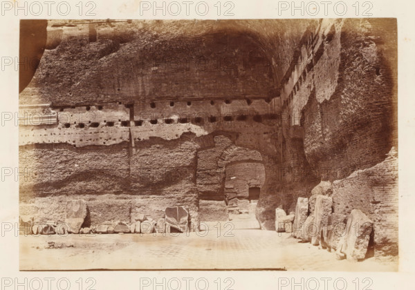 Ruins of the Baths of Caracalla on the Aventine Hill in Rome, photograph, page, Aventine, c. 1863 - in or before 1873, photographer, Rome, paper, albumen print, height 100 mm x width 152 mm, ruin of a building, architecture, public baths, Baths of Caracalla, A sepia-toned photograph showing a large, rough stone wall with visible layers and holes, featuring an arched opening in the center leading into a darker recess, rubble and broken stone blocks arranged along the base, a row of larger stone fragments and carved pieces lined on the ground to the left and right, textured vertical surfaces on the right side, and a patterned ground area in the foreground.
