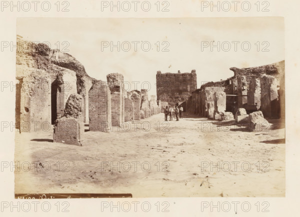 Ruins at Ostia Antica, photograph, page, Ostia Antica, c. 1863 - in or before 1873, photographer, Ostia, paper, albumen print, height 101 mm x width 150 mm, Photo after page 92., ruin of a building, architecture, A wide sandy pathway runs through a scene of weathered stone and brick ruins with tall rectangular columns and partial walls on both sides, some surfaces rough and crumbled while others show layered brickwork; a large square tower stands near the center background with irregular top edges, and low earthen mounds and broken masonry pieces are scattered along the ground; a small group of people in period clothing stands near the middle of the path, and the sky above is pale and mostly clear.