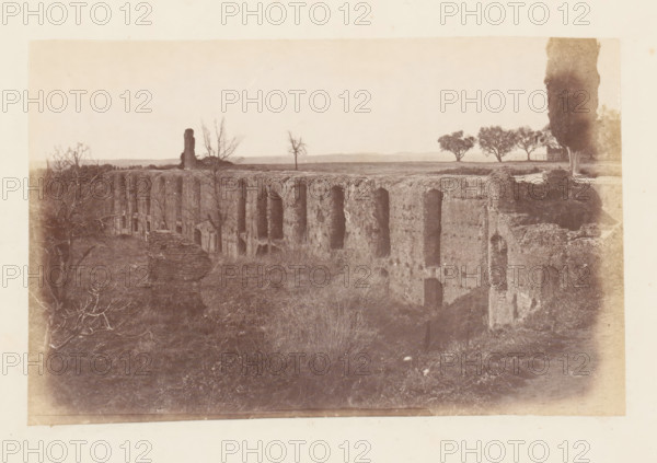 View of the ruins of the Villa Adriana at Tivoli, photograph, page, Tivoli, c. 1863 - in or before 1873, photographer, paper, albumen print, height 95 mm x width 153 mm, villa, ruin of a building, architecture, Villa Adriana, A wide sepia-toned photograph showing a long, weathered stone structure with a series of tall arch-shaped openings along its vertical face, set in an open grassy area. In front of the structure there is overgrown vegetation and low shrubs. Leafless tree branches appear on the left foreground, and a lone bare tree stands on the flat top of the structure. On the right side a tall, narrow tree rises above the ground near a small fence and a cluster of fuller trees further back. The horizon is low and distant, with a pale sky occupying most of the upper portion of the image.