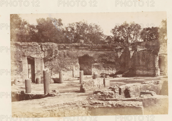View of the ruins of the Villa Adriana at Tivoli, photograph, page, Tivoli, c. 1863 - in or before 1873, photographer, paper, albumen print, height 100 mm x width 153 mm, villa, ruin of a building, architecture, Villa Adriana, A sepia-toned photograph of ancient stone ruins with weathered brick walls and arched openings, scattered column fragments standing upright with fluted surfaces and broken tops, low rubble and rectangular masonry blocks across a rough foreground, a large recessed archway in the central wall, vertical wall recesses at the edges, and a line of trees and foliage visible above the ruin wall against a pale sky.
