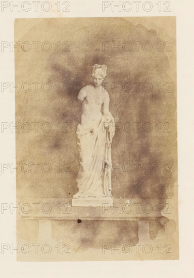 Photo reproduction of a painting, depicting a sculpture of Venus, photograph, reproduction, page, after painting by anonymous, c. 1863 - in or before 1873, photographer, paper, albumen print, height 151 mm x width 98 mm, sculpture, bath of Venus, A pale, weathered photograph shows a classical marble statue of a draped female figure standing on a rectangular plinth against a mottled, darkened background; the figure has an upstyled hair arrangement and a partially exposed torso with drapery wrapped around the lower body and falling in soft folds, one arm missing at the shoulder while the other holds a gathered portion of the cloth, and the image has uneven discoloration and faint stains around the edges of the paper.