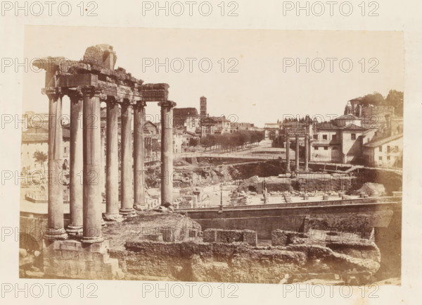 View of the Roman Forum in Rome, with the ruins of the Temple of Saturn in the foreground, photograph, page, Roman Forum, c. 1863 - in or before 1873, photographer, Rome, paper, albumen print, height 100 mm x width 153 mm, landscape with ruins, temple, shrine, Roman religion, Temple of Saturn, A sepia-toned photograph showing tall classical columns with ornate capitals and a partially ruined entablature in the foreground, standing on a stone base above excavated ruins and stone foundations. In the middle distance there are additional isolated columns and fragments scattered among open trenches and low walls. A paved roadway or promenade lined with trees runs behind the ruins, with a mix of masonry buildings, a tower, and tiled-roof structures along its edge. The sky appears pale and empty above the scene, and the image has a wide margin of lighter paper around the printed area.
