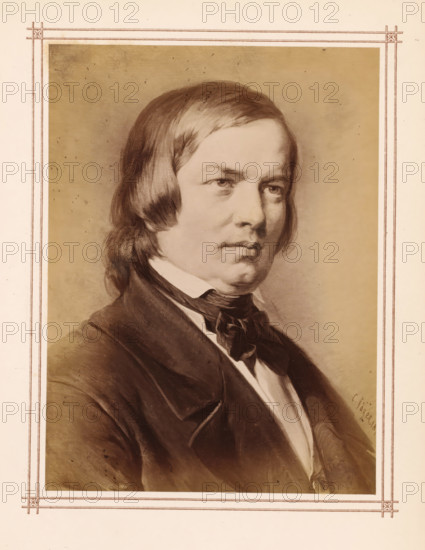 Photo reproduction of a portrait of Robert Schumann by Carl Jaeger, photograph, reproduction, page, name: 'C. Jäger 18...', after painting by Carl Jaeger, c. 1865 - c. 1875, photographer, paper, albumen print, height 144 mm x width 106 mm, Person, Robert Schumann, composer, A sepia-toned portrait of a man shown from the chest up, with medium-length straight hair swept to the sides, a serious facial expression, and visible eyes looking slightly off center, wearing a dark jacket over a white shirt and a dark tied scarf at the collar, set against a plain shaded background and framed by a decorative double-lined border.