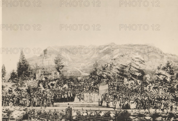 Festakt. Schlussbild, View of the final performance of the Calven-Feier in Chur, page, photomechanical print, number: 'V.', photographer: Lienhard & Salzborn, Chur, c. 1898 - in or before 1899, paper, collotype, photoprint, height 180 mm x width 254 mm, group of actors, troupe, actors on the stage, mountains, A wide sepia-toned scene shows a large outdoor gathering set against a backdrop of rocky mountains and trees, with a multitude of people assembled on tiered stone steps and rocky outcrops. Many individuals wear varied historical costumes and garments, including long dresses, cloaks, and headwear, and several people hold large flags and banners. In the foreground a group of musicians with instruments sit behind a low barrier, and in front of them performers stand on a raised platform where a few figures with distinctive robes and shields are positioned. To the sides and behind the platform the crowd rises densely, interspersed with tall evergreen trees, rocky formations, and what a...