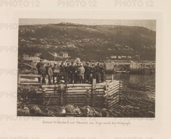 Group portrait of Emperor Wilhelm II of Germany and his entourage at Skårøya, Kaiser Wilhelm II auf Skaarö, at the age of Waljagd., page, photomechanical print, photographer: Paul Güssfeldt (possibly), Norway, cliché maker: Reichsdruckerei, publisher: Paetel, Gebrüder, Berlin, c. 1889 - in or before 1892, paper, photoaquatint, height 109 mm x width 151 mm, Person, Wilhelm II (Emperor of Germany), emperor, anonymous historical persons depicted in a group, in a group-portrait, A sepia-toned photograph shows a group of people standing on a wooden dock structure made of stacked logs at a rocky shoreline, with several small wooden buildings and elevated walkways visible behind them and a hillside rising in the background; a small boat rests by the dock and people on the dock wear a mix of dark and light clothing and hats, while rocks and water are visible in the foreground.