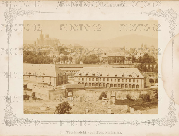 View of Metz, Total view of Fort Steinmetz, Metz and Seine Umgebung, photograph, page, number: '1.', photographer: C. Holt, Metz, publisher: Verlag der Deutschen Buchhandlung, c. 1865 - in or before 1875, paper, albumen print, albumendruk, height 99 mm x width 141 mm, Photo opposite title page., city-view, and landscape with man-made constructions, A sepia-toned panoramic view framed by decorative borders shows a townscape with a large multi-story building in the foreground featuring a row of arched openings on the ground level and dormer windows on a sloped roof, set behind a fortified wall with an arched gateway. Behind this complex are rows of trees and additional buildings with varied roofs and architectural shapes, and further back a dense cluster of houses and taller structures including a cathedral-like building with towers and steep vertical elements. Hills rise faintly in the distant background under a pale sky, and the entire scene has an aged, antique photograph appearance.