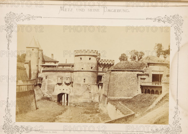 Das Deutsche Thor, View of the Porte des Allemands in Metz, Metz und seine Umgebung, photograph, page, number: '5.', photographer: C. Holt, Metz, publisher: Verlag der Deutschen Buchhandlung, c. 1865 - in or before 1875, paper, albumen print, albumendruk, height 99 mm x width 155 mm, city-gate, Porte des Allemands, A sepia-toned photograph shows a fortified stone structure with rounded towers and battlements, connected by thick walls and a bridge-like entrance over a narrow water channel, flanked by sloped earthworks and grassy embankments. The left side features tall conical-roofed towers and attached buildings with tiled roofs, while the central section displays a cylindrical keep with small window openings and crenellations. The right side includes a carved stone façade with an open balcony above arched openings, and a grassy mound with scattered trees rising behind the fortifications. Decorative ornate borders frame the photograph on all sides.