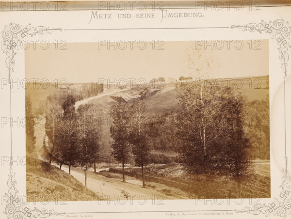 Schlucht von Gravelotte I, View of the gorge near Metz where the Battle of Gravelotte took place in 1870, Metz und seine Umgebung, photograph, page, number: '29.', photographer: C. Holt, Gravelotte, publisher: Verlag der Deutschen Buchhandlung, Metz, c. 1865 - in or before 1875, paper, albumen print, albumendruk, height 99 mm x width 153 mm, dale, valley, A sepia-toned photograph framed by an ornate decorative border shows a rural landscape with a dirt road winding beside a row of tall trees, a sloped embankment and a low stone wall alongside the road, open fields and gently rolling hills in the background, clusters of trees and a stand of taller trees further along the valley, and a sparse foreground of grasses and bushes.