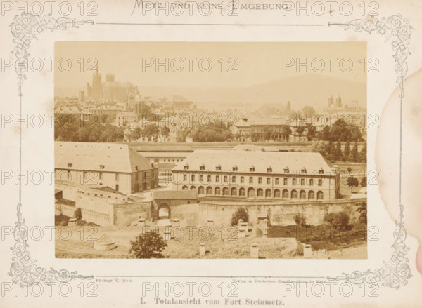Metz und seine Umgebung, book, 1875, textile materials, pasteboard, paper, linen (material), albumen print, color on cut, height 210 mm x width 160 mm x thickness 20 mm, A sepia-toned photograph centered within a decorative border shows a panoramic urban scene with a large multi-story building in the foreground featuring a long row of arches along its ground level and steeply pitched roofs with small dormer windows, adjacent to another rectangular building with a sloped roof and small windows. A fortified wall with an arched gateway runs beneath these buildings, and open ground with scattered low structures and a small circular roofed feature lies in front. Behind the foreground complex, rows of trees and varied residential and institutional buildings fill the middle distance, with a prominent cathedral-like structure rising above the city skyline to the left and additional church towers visible toward the right, all set against hazy rolling hills in the background. The image is framed...