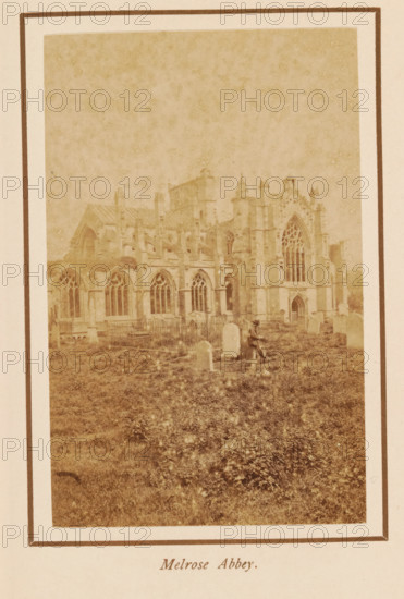 Melrose Abbey, photograph, page, photographer: George Washington Wilson, Scotland, c. 1885 - c. 1890, paper, albumen print, height 108 mm x width 68 mm, ruin of church, monastery, Melrose (Scotland), A faded sepia photograph shows the ruins of a large Gothic stone abbey with tall arched windows and detailed tracery, buttresses, and a prominent ornate façade; the building sits behind a grassy area with scattered upright tombstones and a fenced section of graves, and a solitary seated figure wearing a hat is visible among the stones near the front of the abbey.