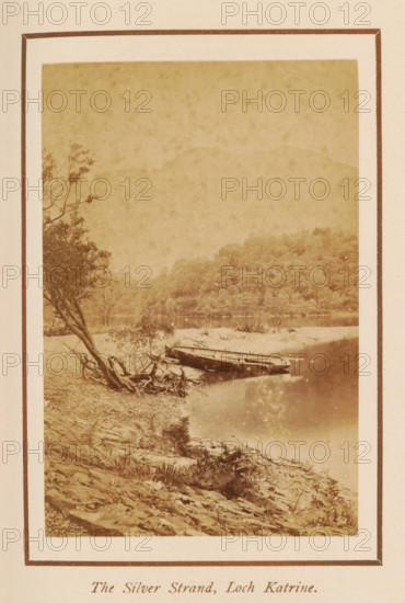 The Silver Strand, Loch Katrine, photograph, page, photographer: George Washington Wilson, Scotland, c. 1885 - c. 1890, paper, albumen print, height 104 mm x width 69 mm, lake, ships, boats on land, beached, A sepia-toned photograph bordered by a double mat shows a calm shoreline scene with a gently sloping rocky bank in the foreground, a fallen tree with exposed roots leaning toward the water on the left, and a small wooden boat pulled up at the water's edge. The water reflects light and the opposite shore, where a densely wooded hillside rises, and faint tree branches frame the left side of the image. The overall image has a textured, aged appearance.