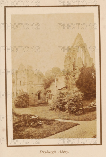 Dryburgh Abbey, photograph, page, photographer: George Washington Wilson, Scotland, c. 1885 - c. 1890, paper, albumen print, height 103 mm x width 69 mm, ruin of church, monastery, A faded sepia photograph framed by a wide margin shows the ruins of a stone abbey with tall, crumbling arches and window openings, surrounded by dense foliage and bushes; a grassy foreground with a curving path leads toward a low stone wall and scattered stones, and trees rise behind and around the ruined structure.
