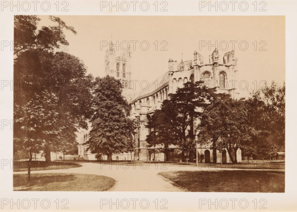 The Cathedral, Lady's Chapel, View of Canterbury Cathedral, photograph, page, number: '25673', Canterbury Cathedral, c. 1882 - in or before 1892, photographer, Kent, Canterbury, paper, albumen print, height 113 mm x width 174 mm, church (exterior), parts of church exterior and annexes: chapel, A sepia-toned photograph showing a large stone building with gothic architectural features partially obscured by tall leafy trees, a curved pathway in the foreground bordered by grassy areas, arched windows and buttresses visible on the building, a tall tower rising behind the trees, and scattered smaller trees and shrubbery around the structure.