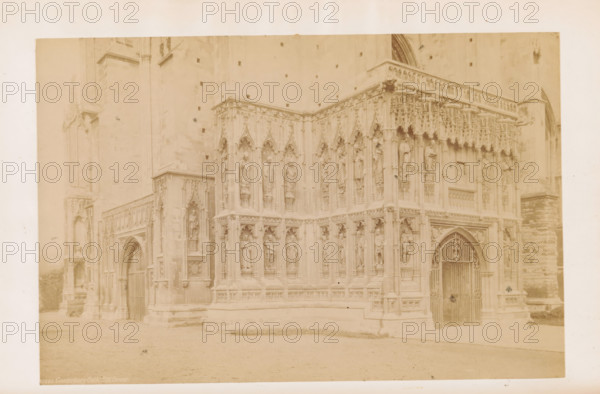 The Cathedral, Southwest Corner, View of Canterbury Cathedral, photograph, page, Canterbury Cathedral, c. 1882 - in or before 1892, photographer, Kent, Canterbury, paper, albumen print, height 115 mm x width 174 mm, church (exterior), parts of church exterior and annexes: portal, A faded sepia photograph showing a stone building corner with ornate Gothic carvings and sculpted figures set in niches across two levels, pointed arches, and decorative tracery; a gated arched doorway with an iron gate is visible on the right side, flanked by carved columns and statues; the left side shows another arched entrance partially recessed beneath detailed stonework; the ground in front appears smooth and level, and the upper facade shows small square openings and a plain stone wall above the carved section.