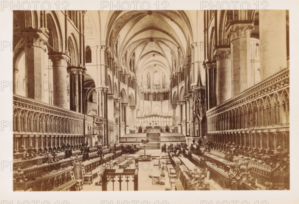 The East Choir, Interior of Canterbury Cathedral with a view of the choir, photograph, page, Canterbury Cathedral, c. 1882 - in or before 1892, photographer, Kent, Canterbury, paper, albumen print, height 115 mm x width 175 mm, parts of church interior: choir, A wide interior view of a large cathedral nave with tall vaulted ceilings and pointed arches, flanked by rows of ornate wooden choir stalls with carved details and high backs on both sides, a central aisle leading toward an elevated chancel area with steps and an ornate screen, massive stone columns with decorated capitals supporting the arches, slender clerestory windows high above, decorative stone tracery and carved ornament along the walls, scattered chairs and a few candlesticks or stands along the aisle, and architectural details extending symmetrically toward a distant apse.