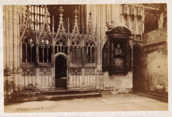Interior of a transept of Canterbury Cathedral, The Transept of the Martyrdom, photograph, page, number: '25689', Canterbury Cathedral, c. 1882 - in or before 1892, photographer, Kent, Canterbury, paper, albumen print, height 115 mm x width 175 mm, parts of church interior: transept, A sepia-toned photograph of an ornate stone interior showing a series of pointed Gothic arches with tracery and tall narrow windows, ornate finials and carved vertical ribs, and a central arched doorway leading into a darker interior. To the right of the arches a sculpted wall monument sits within a recessed niche framed by columns and an arched pediment, featuring a carved bust set above a rectangular plaque. The surrounding walls are composed of dressed stone blocks, with a secondary arched passageway visible on the far right and worn stone steps and a tiled floor in the foreground.