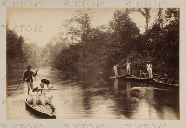 Crocodile hunting party, album page, photograph, This photo is part of an album., number: '56', photographer, Sumatra, c. 1890 - c. 1900, paper, pasteboard, albumen print, height 265 mm x width 355 mm, In the list of images, this photo is described with the words 'Flusspartie in Bedagei'., river (landscape with figures, staffage), crocodiles: crocodile, hunt, other animals, A sepia-toned photograph showing a calm river bordered by dense, leafy tropical vegetation and tall trees, with several wooden narrow boats on the water. In the foreground boat, a standing man holds a long pole, while two seated men in light clothing and hats face forward, one pointing and the other holding a paddle. Further back, another boat holds a standing man with a pole, a man standing with hands near his head, and a seated figure, while a third boat nearby contains two seated shirtless men. The water shows gentle ripples and reflections of the trees, and the sky above is light and unobstructed.