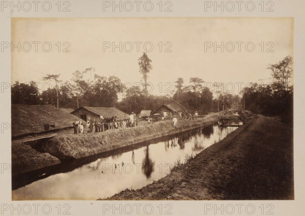 Canal at the Stabat plantation, Sumatra, album page, photograph, This photo is part of an album., number: '9', photographer, c. 1890 - c. 1900, paper, pasteboard, albumen print, height 243 mm x width 372 mm, In the list of images, this photo is described with the words 'Kanal in Stabat Estate'., canal, plantation, working class, labourers, A sepia-toned photograph showing a narrow waterway reflecting trees and the sky, with earthen banks on both sides and a dirt path along the near bank, a group of people standing along the far bank near thatched-roof huts and simple wooden structures, a few carts and a person seated on one of them, clusters of trees rising behind the huts including a tall solitary tree, a small wooden footbridge crossing the water farther down, and a person seated on the bridge while another stands nearby.