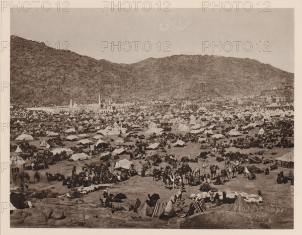 Pilgrim camp in Mina near Mecca during the Hajj, Westlicher Theil des sich von Westen nach Osten erstreckenden Thales Muna (Mina) während der grossen jährlichen Pilgerversammlung, album page, photomechanical print, This print is part of an album., number: 'X', photographer: Abd al-Ghaffar, Mina, cliché maker: anonymous, 1889, Saudi Arabia, pasteboard, paper, collotype, photoprint, height 270 mm x width 360 mm, tent-camp, Hajj, Period, 1888-03-30 - 1888-04-27, A wide landscape shows a crowded encampment spread across a flat area at the base of rocky hills, with numerous tents of various shapes and sizes clustered closely together and rows of low buildings along the edge of the settlement; many people are scattered among the tents, some standing, some seated, and groups are gathered around camels and packs, while camels lie or stand throughout the foreground; the scene is monochrome and framed by a pale border, with rugged hills rising behind the settlement and a clear sky above.