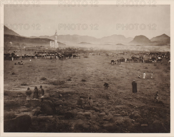 Die zwischen Muna und Arafah situated Pilgerstation Muzdal'fah, View of Muzdalifah, located between Mina and the plain of Arafat, during the Hajj, album page, photomechanical print, This print is part of an album., number: 'XII', photographer: Abd al-Ghaffar, Muzdalifah, cliché maker: anonymous, 1889, Abd al-Ghaffar, photographer, cliché maker, Saudi Arabia, pasteboard, paper, collotype, photoprint, height 270 mm x width 360 mm, other ritual practices, Islam, Mohammedanism, Hajj, Period, 1888-03-30 - 1888-04-27, A wide, sepia-toned landscape shows a flat, rocky plain stretching toward distant hazy mountains under a pale sky, with a tall tower and adjacent building near the middle left skyline. Groups of people are scattered across the plain, some standing, some sitting, and several small clusters of animals, including pack animals, are gathered near the center and right. In the foreground three figures stand together facing away, and a few other solitary figures sit or stand on rocky g...