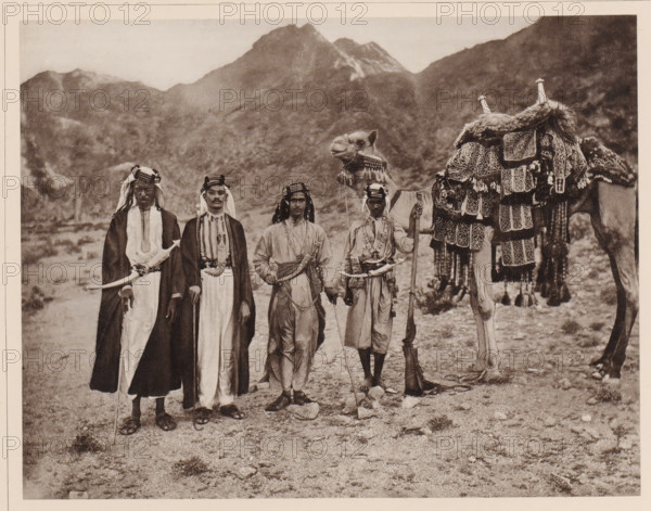 Das Reitkamel (Hèdjin) des Scherif Jahja, eines Sohnes des Scherif Ahmed, dessen Vater der beruhmte 1886 verstorbene Grossscherif Abd èl-Muttálib with a grain of silver gestickten Satteldecke (batat), the Zaum hält der Sklave Jahja's, neben diesem steht Jahja selbst im Reiteranzug and on the left in long Kleideren zwei Scherife niedrigeren Ranges, Group portrait of sjarif Jahja with on the right an enslaved boy with a dromedary and on the left two unknown sjariffs, album page, photomechanical print, This print is part of an album., number: 'XVII', photographer: Abd al-Ghaffar, Saudi Arabia, cliché maker: anonymous, 1889, Abd al-Ghaffar, photographer, cliché maker, pasteboard, paper, collotype, photoprint, height 270 mm x width 360 mm, Person, Jahja, historical persons, historical persons not known by name, hoofed animals: dromedary, family of the sovereign, Four men stand side by side in traditional robes and head coverings, each holding a staff or weapon, with one man wearing a long d...