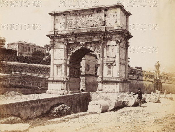 Man posing by the Arch of Titus at the Roman Forum in Rome, photograph, Part of a French artist's album., Rome, c. 1880 - c. 1900, photographer, paper, albumen print, height 194 mm x width 254 mm, triumphal arch, Roman Forum, Arch of Titus, A large stone triumphal arch with a central arched opening stands prominently in the scene, showing carved reliefs, fluted columns, and weathered surfaces with visible cracks and dark stains; fragments of broken columns and cylindrical stone drums lie on the ground in front of the arch; a man wearing a dark coat and hat stands beside the stone fragments; a low stone wall and walkway run alongside the arch and foreground; behind and to the left of the arch there is a multi-windowed building partially obscured by trees and shrubbery; to the right a long wall with decorative statues and architectural details extends into the distance; the overall image has a sepia tone and shows strong contrasts between the stone structures and the lighter sky.