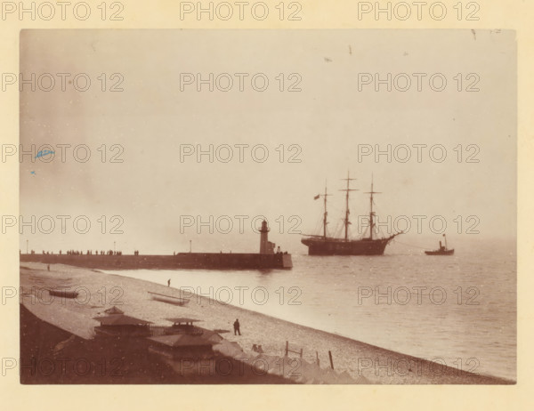 Tugboat takes sailing ship in tow at a harbor pier, photograph, Part of a French artist's album., c. 1880 - c. 1900, photographer, pasteboard, paper, albumen print, albumen print on cardboard, height 119 mm x width 166 mm, sailing-ship, sailing-boat, pier, quay, wharf, tug, pusher tug, A faded seaside scene shows a pebble beach in the foreground with small boats resting on the shore and a solitary figure standing near the water, a line of beach huts and low structures along the edge, a long stone pier extending into the water with a small lighthouse at its end and silhouettes of people standing along the pier, a large sailing ship with tall masts anchored offshore and a smaller steam-powered vessel nearby, calm water reflecting the vessels, and a pale sky above with some scattered spots and imperfections on the photograph.