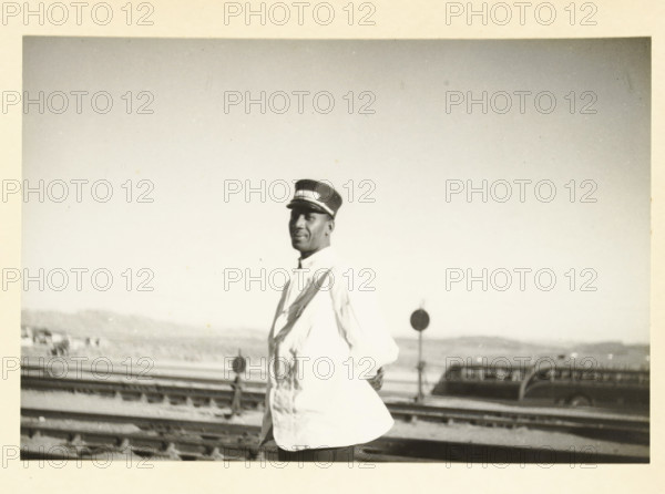 Portrait of an American man, probably working on a train, Unknown location in the United States: portrait of a black man (probably working on a train) (former title), My Negro, photograph, title: 'MY NEGRO.', photographer: attributed to Wouter Cool, United States of America, 1936, North America, Attributed to, pasteboard, paper, gelatin silver print, height 150 mm x width 222 mm, Americans with African roots, African-Americans, African Americans, A black and white photograph showing a man standing near railroad tracks wearing a light-colored jacket and a peaked cap with a contrasting band, his hands behind his back and his face turned slightly toward the camera, with distant hills and blurred structures in the background and a round sign visible near the tracks.