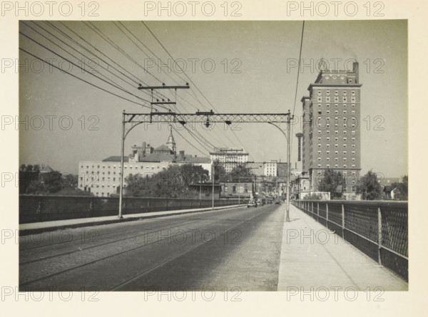 Unknown location in the United States: view in a city, Entrance of a medium-sized American city, photograph, title: 'ENTRANCE OF A MID-SIZED AMERICAN CITY', photographer: attributed to Wouter Cool, United States of America, 1936, North America, Attributed to, pasteboard, paper, gelatin silver print, developing gelatin silver print, height 157 mm x width 225 mm, height 315 mm x width 285 mm, city-view, and landscape with man-made constructions, A wide street or bridge with tram rails runs toward a distant urban area, flanked on the right by a sidewalk and a metal railing and on the left by a low barrier. Overhead metal trusses and poles span the roadway, supporting multiple parallel wires that angle across the sky. Near the horizon there are several buildings including a tall multi-story brick building with many evenly spaced windows and a decorative roofline, lower commercial and institutional buildings with varied roof shapes, and a structure with a pointed spire. A few small vehicles...