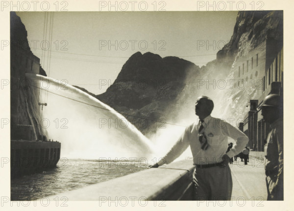 Water outlet from a power plant at an unknown location in the United States, Water outlet 60m. fall height, photograph, title: 'WATER OUTLET, 60m. FALL HEIGHT', photographer: attributed to Wouter Cool, United States of America, 1936, North America, Attributed to, pasteboard, paper, gelatin silver print, developing gelatin silver print, height 157 mm x width 225 mm, height 315 mm x width 272 mm, power station, A black and white photograph showing a large curved stream of water arcing from the left toward the center, creating mist against a backdrop of rugged dark mountains. In the foreground a man in a white shirt and patterned tie leans on a railing, his face turned upward and his hand resting on his hip, while another figure wearing a helmet stands to the right along a paved walkway with a building facade behind them. The left side shows a concrete structure with vertical elements near the water and thin cables stretching across the sky above the scene.