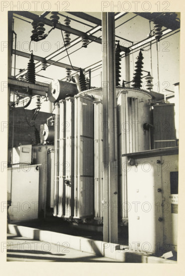 Transformer at an Electric Power Station, United States, photograph, title: '132 kV TRAFO.', photographer: attributed to Wouter Cool, United States of America, 1936, North America, Attributed to, pasteboard, paper, gelatin silver print, developing gelatin silver print, height 230 mm x width 156 mm, height 315 mm x width 330 mm, power station, A black-and-white photograph showing a section of electrical equipment with tall cylindrical and rectangular metal components, ribbed cooling fins, and large insulators mounted on a metal framework. Overhead metal beams and crossbars support suspension insulators and heavy cables that run between the structures. A vertical metal column stands in the foreground partially obscuring the equipment behind it. Shadows fall across the concrete base and the side of an enclosed cabinet with visible hinges and fasteners. The scene has a high-contrast, slightly grainy appearance.