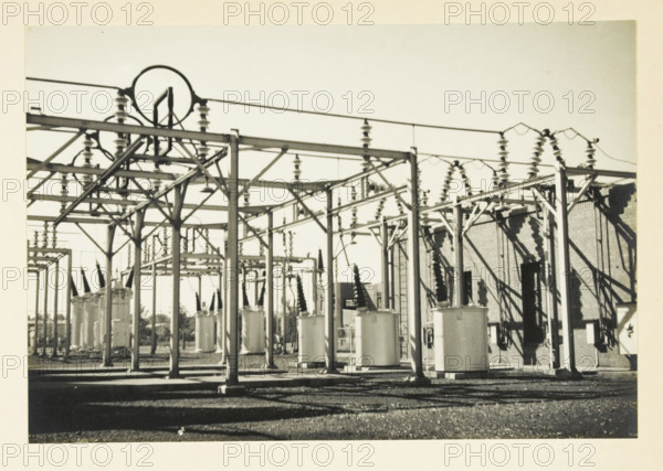 Switch installation of current-voltage transformers and isolating switches, Transformers and switches at an unknown location in the United States, photograph, title: 'SWITCH INSTALLATION OF CURRENT-VOLTAGE TRANSFORMERS, AND ISSUING SWITCHES', photographer: attributed to Wouter Cool, United States of America, 1936, North America, Attributed to, pasteboard, paper, gelatin silver print, developing gelatin silver print, height 155 mm x width 227 mm, height 315 mm x width 330 mm, power station, A wide outdoor industrial area with a grid of metal frameworks, insulators, and cylindrical tanks beneath overhead wires, with shadows cast on a gravel ground and a brick building partially visible at the right.