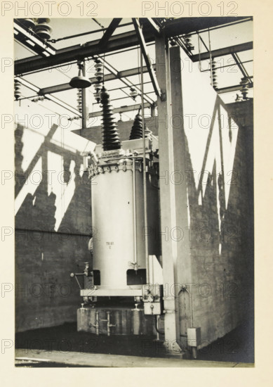 Oil switch in a power station at an unknown location in the United States, 132 kv oil switch, photograph, title: '132 kV OIL SWITCH.', photographer: attributed to Wouter Cool, United States of America, 1936, North America, Attributed to, pasteboard, paper, gelatin silver print, developing gelatin silver print, height 225 mm x width 155 mm, height 315 mm x width 330 mm, power station, A black and white photograph of an industrial interior showing a large cylindrical transformer-like device mounted on a base with insulating bushings and coiled connectors at the top, set between vertical structural columns and surrounded by overhead metal framework and insulators; strong diagonal shadows fall across the concrete walls behind the equipment, and various pipes, brackets, and mounting hardware are visible along the column and base.