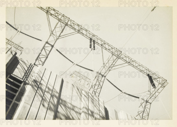 Detail of a power plant at an unknown location in the United States, 280 kv input at the power plant, photograph, title: '280 kV INPUT, AT THE POWER PLANT', photographer: attributed to Wouter Cool, United States of America, 1936, North America, Attributed to, pasteboard, paper, gelatin silver print, developing gelatin silver print, height 154 mm x width 227 mm, height 315 mm x width 272 mm, power station, A low-angle view shows a lattice steel framework of electrical transmission structures towering above a concrete surface with vertical grooves and stair-like shadowed openings, suspended insulator strings and curved cables drape between the metal towers, coiled cylindrical components hang from a crossbeam, and long shadows of the framework fall across the concrete.