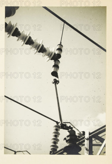 Protection of the voltage transformer, Protection of a transformer in an electrical wire, photograph, title: 'PROTECTION OF THE, VOLTAGE TRANSFORMER.', photographer: attributed to Wouter Cool, United States of America, 1936, North America, Attributed to, pasteboard, paper, gelatin silver print, height 228 mm x width 155 mm, height 315 mm x width 330 mm, pylons, power lines, A close-up view looking upward at electrical insulators and hardware against a pale sky, showing a diagonal string of disc-shaped insulators connected to a metal rod and suspended components, cylindrical insulator elements hanging from a thin suspension, metal brackets and crossarms with attached fittings, and multiple straight wires and structural beams crossing the frame.