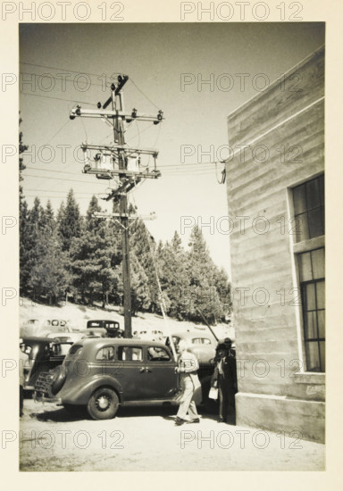 One of the famous masts but a beautiful one, Electricity pylon at an unknown location in the United States, photograph, title: 'ONE OF THE FAMOUS MATTERS, BUT A BEAUTIFUL ONE', photographer: attributed to Wouter Cool, United States of America, 1936, North America, Attributed to, pasteboard, paper, gelatin silver print, height 229 mm x width 156 mm, height 315 mm x width 215 mm, pylons, power lines, A black and white photograph showing a tall wooden utility pole with crossarms and multiple insulators and wires against a clear sky, a grouped backdrop of evergreen trees on a hillside, a lineup of vintage cars parked along the road with one car prominent in the foreground, and two men near the building edge—one walking past the car and another standing against the building holding a small bag. The right side of the image shows the textured exterior wall of a building with a tall window.
