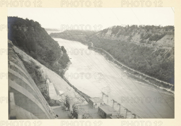 Niagara Falls Cataracts, Niagara Falls, United States, Canada, photograph, title: 'NIAGARA FALLS, CATARACTS.', photographer: attributed to Wouter Cool, United States of America, 1936, North America, Attributed to, pasteboard, paper, gelatin silver print, developing gelatin silver print, height 152 mm x width 226 mm, height 315 mm x width 285 mm, waterfall, A grayscale photograph showing a wide river flowing between steep, tree-covered cliffs with layered rock faces; the near left side includes a curved concrete structure descending toward the water and a sloped embankment, while the lower foreground shows several rectangular industrial buildings and electrical pylons with power lines extending diagonally across the image; the opposite riverbank displays terraces of vegetation and exposed rock, and the distant background fades into a lighter horizon.