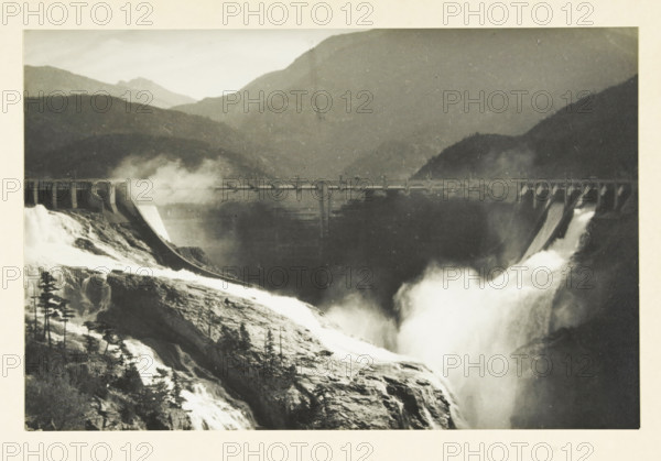 Diablo Dam and Skagit River, Washington state, United States, Skagit River project Diabolo dam, photograph, title: 'SKAGIT RIVER PROJECT, DIABOLO DAM.', photographer: attributed to Wouter Cool, United States of America, 1936, North America, Attributed to, pasteboard, paper, gelatin silver print, developing gelatin silver print, height 150 mm x width 229 mm, height 315 mm x width 330 mm, dam (in canal), A black-and-white photograph showing a large concrete dam spanning a river valley with water rushing over spillways and producing mist; rugged rocky slopes and scattered trees are visible in the foreground leading down to the turbulent water, while layered mountain ridges rise in the background behind the dam.