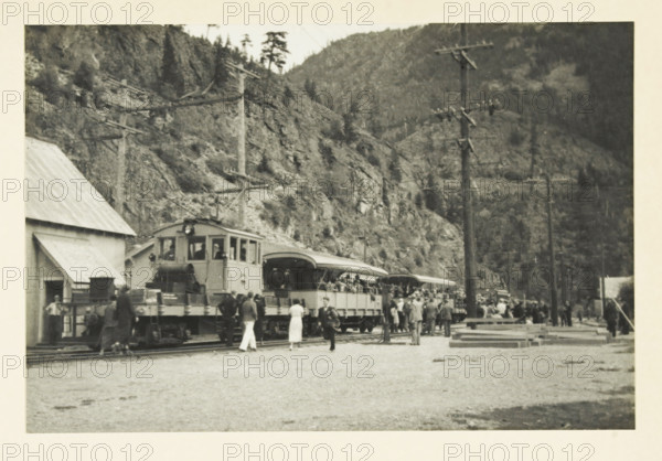Mountain train in the valley of the Skagit River, Washington state, United States, Mountain railway through the valley of the Skagit river Seattle, photograph, title: 'MOUNTAIN STREET THROUGH THE VALLEY OF THE SKAGET RIVER SEATTLE', photographer: attributed to Wouter Cool, United States of America, 1936, North America, Attributed to, pasteboard, paper, gelatin silver print, height 152 mm x width 227 mm, height 315 mm x width 285 mm, passenger train, A black and white photograph showing a train stopped along tracks with an open-sided passenger car and a locomotive at the front, a group of people standing and milling near the train, some dressed in light clothing and some in darker coats, a building with a sloped roof to the left, telegraph poles with crossarms rising beside the tracks, wooden planks and other materials stacked near the right side of the scene, and steep tree-covered rocky hills forming the backdrop.