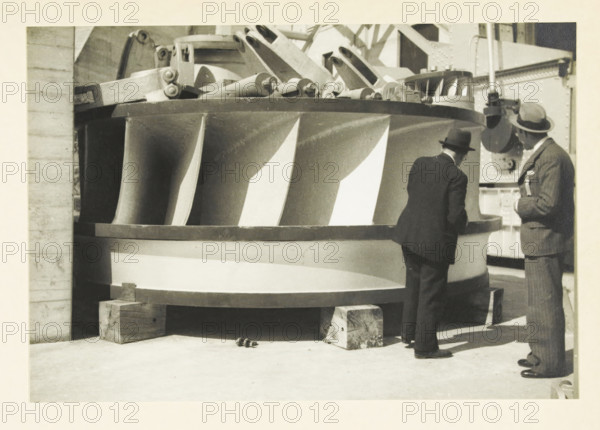 Ariel Dam, Washington state, United States: rotor of the Francis turbine, Rotor of the Francis turbine of Ariel Dam power plant, photograph, title: 'ROTOR OF THE FRANCIS TURBINE, OF ARIEL DAM POWER PLANT', photographer: attributed to Wouter Cool, United States of America, 1936, North America, Attributed to, pasteboard, paper, gelatin silver print, height 153 mm x width 224 mm, height 315 mm x width 285 mm, dam (in canal), A large circular metal structure with curved vertical blades rests on wooden blocks on a concrete floor, showing a patterned surface of alternating light and dark blade faces and a thick rim. Two men in suits and hats stand beside the structure, one facing the rim and leaning slightly forward while the other stands upright holding something near his chest. Behind the structure there are mechanical parts and structural beams with riveted plates and a cylindrical component near the upright man. Small round objects lie on the floor near the base of the structure.