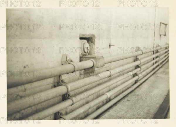 Low-voltage installation on the Ariel Dam, Ariel Dam, Washington state, United States: low-voltage installation, photograph, title: 'LOW-VOLTAGE INSTALLATION, ON THE ARIEL DAM', photographer: attributed to Wouter Cool, United States of America, 1936, North America, Attributed to, pasteboard, paper, gelatin silver print, developing gelatin silver print, height 152 mm x width 222 mm, height 315 mm x width 285 mm, dam (in canal), A grainy black and white photograph of a concrete wall with a row of horizontal metal pipes running along its length, the pipes held by brackets and clamps and appearing worn and slightly rusted, a recessed round fixture set into the wall with a chain hanging from it, a small rectangular box attached near the pipes, and a narrow ledge or walkway below the pipes.
