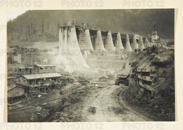 Bonnerville Dam, Portland, Oregon, United States, Portland Oregon, photograph, title: 'BONNERVILLE DAM, PORTLAND-OREGON', photographer: attributed to Wouter Cool, United States of America, 1936, North America, Attributed to, pasteboard, paper, gelatin silver print, developing gelatin silver print, height 152 mm x width 226 mm, height 315 mm x width 330 mm, dam (in canal), A wide black-and-white scene shows a large construction site with a partially built concrete structure made of tall tapered vertical supports and scaffolding, terraces and stepped forms in front of it, and a dirt road running toward the foreground. On the left there are wooden buildings on stilts with pitched roofs and external stairways, and scattered construction materials and equipment around them. A heavy dump truck sits on the dirt road, which is flanked on the right by a steep earthen embankment reinforced with horizontal wooden planks and a group of people standing along the top. In the background a forested hi...