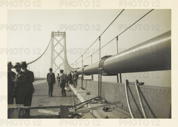 Oakland Bridge, San Francisco, United States, Upper Driving Deck Oakland Bridge, photograph, title: 'Upper Driving Deck OAKLAND BRIDGE, SAN FRANCISCO', photographer: attributed to Wouter Cool, United States of America, 1936, North America, Attributed to, pasteboard, paper, gelatin silver print, developing gelatin silver print, height 154 mm x width 230 mm, height 315 mm x width 272 mm, bridge, A black and white photograph showing a suspension bridge walkway with a large cylindrical cable running along the right side, held by vertical suspenders and wrapped with ropes at intervals, and a tall bridge tower with cross-bracing visible in the background. Several people are standing and walking on the walkway, most wearing hats and coats, with a few clustered near the left edge and others further down the path. The foreground shows part of the railing and a section of the bridge deck with scattered wooden planks and metal fixtures. The sky appears clear and light in tone.