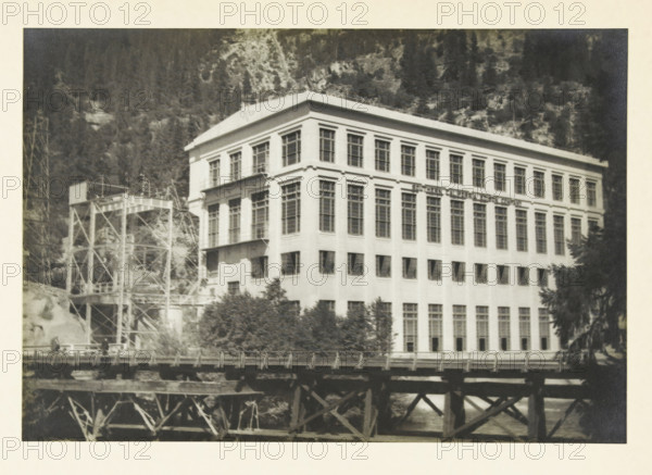 Centrale a, d Big Creek, Fresno California, Big Creek Power Plant, Fresno, California, United States, photograph, title: 'CENTRALE A, D BIG CREEK, FRESNO CALIFORNIE', photographer: attributed to Wouter Cool, United States of America, 1936, North America, Attributed to, pasteboard, paper, gelatin silver print, developing gelatin silver print, height 156 mm x width 228 mm, height 315 mm x width 285 mm, power station, A large multi-story rectangular building with many tall windows occupies the center of the image, its light-colored facade showing rows of uniformly spaced window openings and decorative cornice lines near the roof, and a flat roof. To the left of the building a metal scaffolding or industrial framework with platforms and diagonal supports rises up, partially obscured by trees and vegetation at its base. In front of the building a wooden trestle or bridge structure with visible beams and supports spans horizontally across the lower part of the image, and water can be seen be...