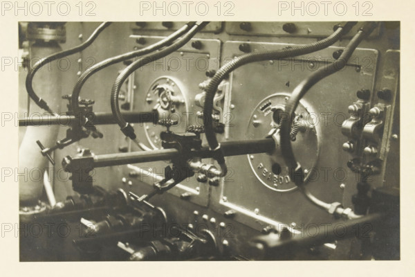 Gas and oil supply under the boilers centrala Long beach Los Angelos, Long Beach, Los Angelos, California, United States: detail of the gas and oil supply of the boilers of the (power) plant, photograph, title: 'GAS AND OIL SUPPLY UNDER THE BOILER, CENTRAL LONG BEACH LOS ANGELOS', photographer: attributed to Wouter Cool, United States of America, 1936, North America, Attributed to, pasteboard, paper, gelatin silver print, developing gelatin silver print, height 141 mm x width 224 mm, height 315 mm x width 330 mm, power station, A close-up view of mechanical equipment showing metal panels with circular access covers and mounted fittings, flexible corrugated hoses and smooth pipes attached to the covers, several valve handles and clamps, rows of bolts and rounded fasteners on the panels, and various connecting rods and fixtures extending horizontally and diagonally across the scene.