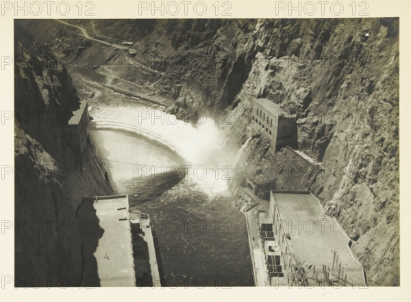 View into the Black Canyon, United States, photograph, title: 'VIEW INTO THE BLACK CANYON', photographer: attributed to Wouter Cool, United States of America, 1936, North America, Attributed to, pasteboard, paper, gelatin silver print, developing gelatin silver print, height 157 mm x width 225 mm, height 315 mm x width 330 mm, ravine, chasm, abyss, canyon, A black and white aerial view of a canyon with steep rocky walls and a curved dam or spillway releasing a large plume of water that splashes against the rock face, adjacent concrete structures with flat roofs and rectangular openings built into the canyon sides, a narrow winding road visible along the upper rim, and turbulent dark water flowing below framed by lighter colored rock and concrete surfaces.